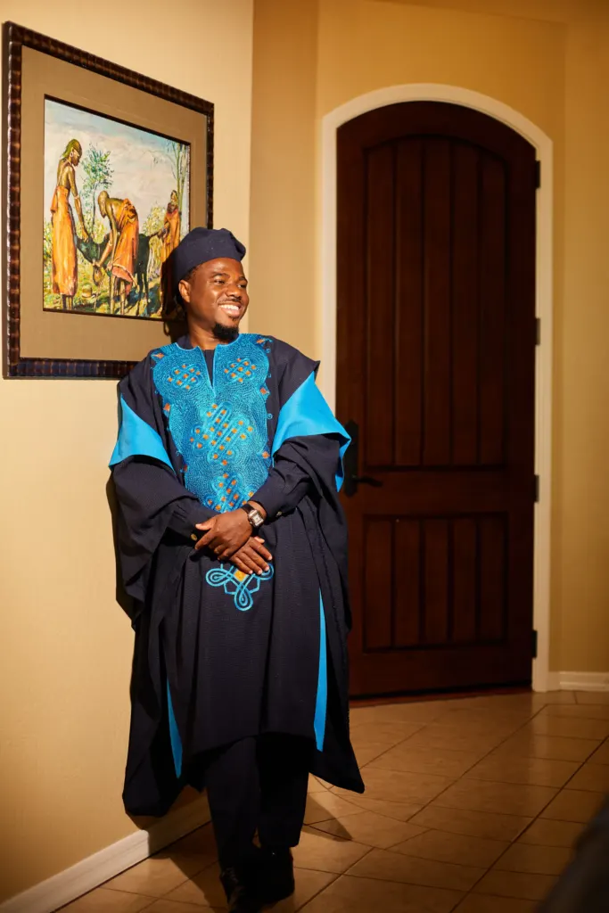 Groom in a flowing navy and blue embroidered robe and cap, laughing in a warm hallway