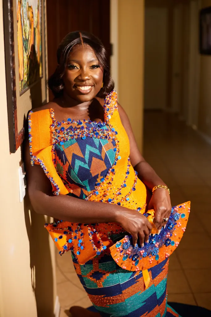 Bride-to-be in a kente gown holding a fan, smiling against a golden hallway wall