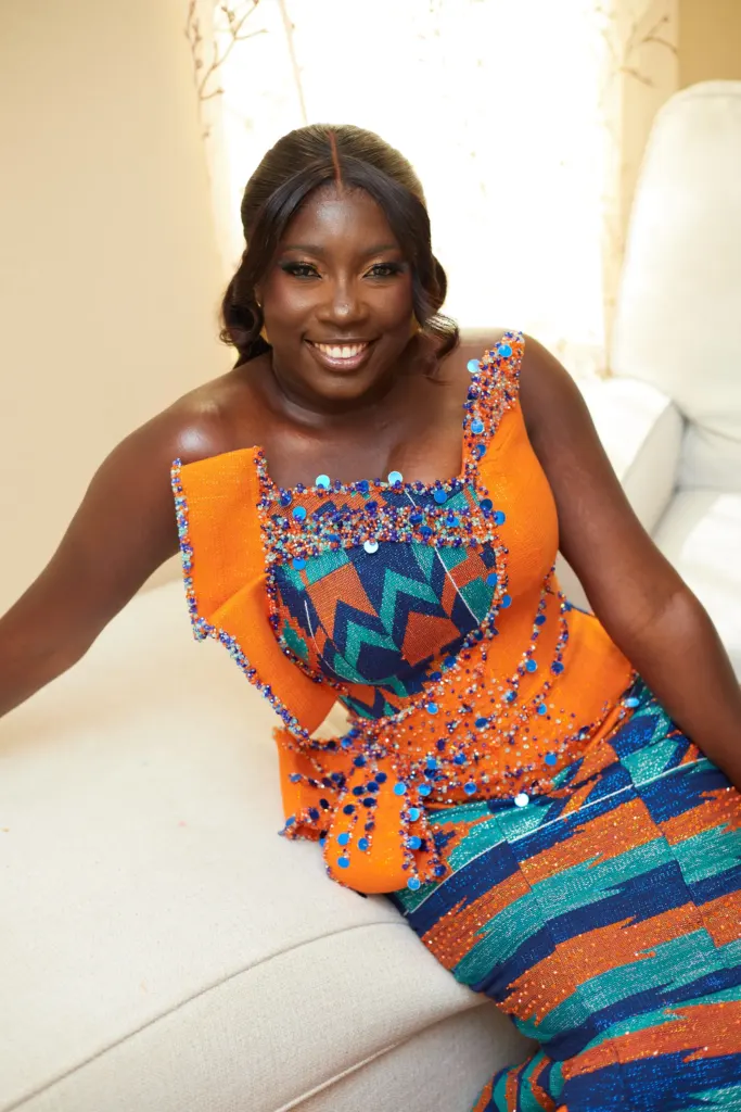 Bride-to-be in an orange and blue kente gown leaning back on a white sofa, smiling