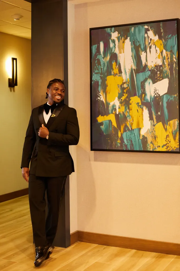 Man in a double-breasted tuxedo beside a large abstract painting, adjusting his cufflink