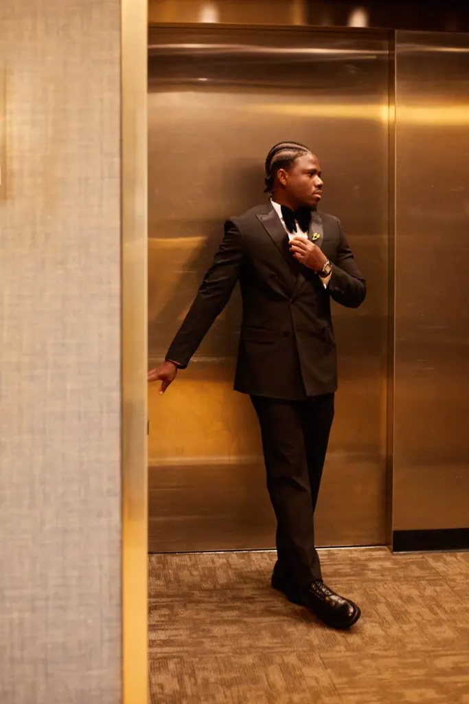 Man in a tuxedo leaning in a golden elevator doorway, straightening his bow tie