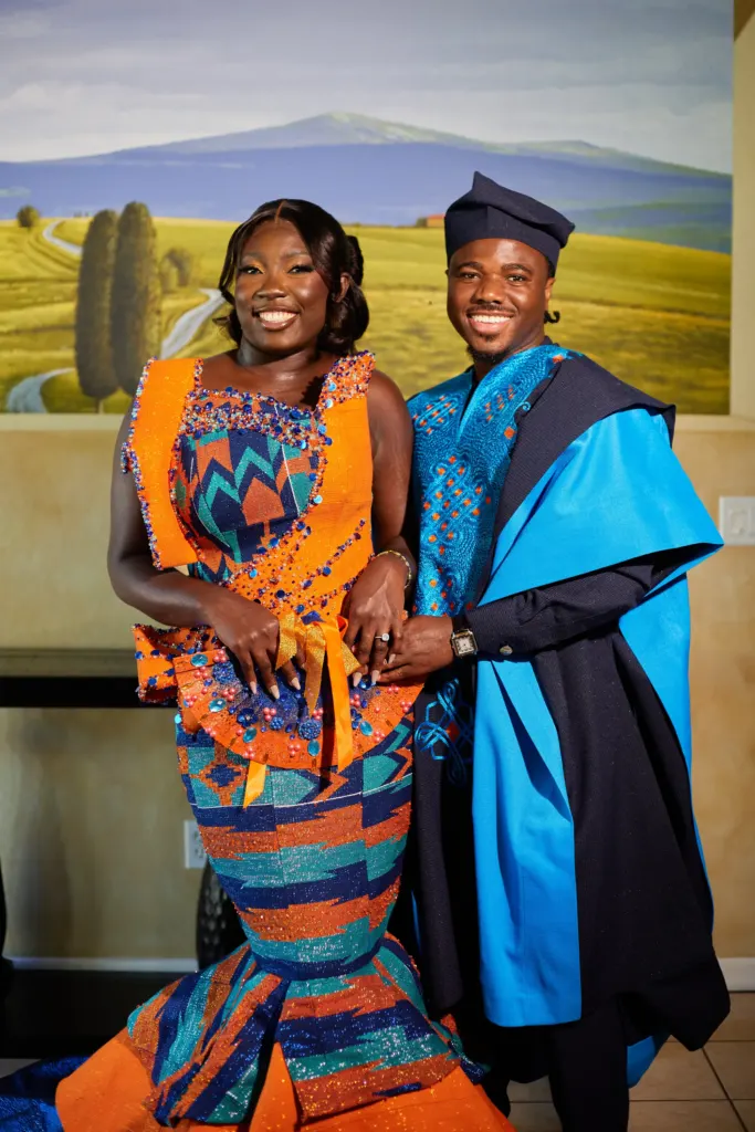 Couple in matching orange kente and blue embroidered robes in front of a painted landscape mural