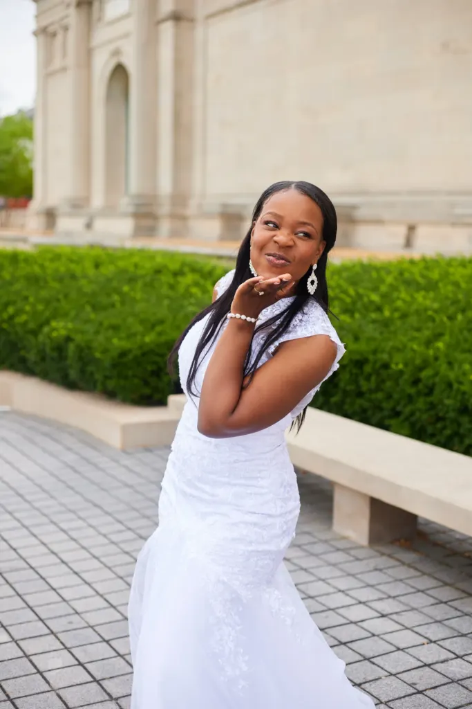 Bride blowing a kiss outside a stone museum, clipped hedges behind her