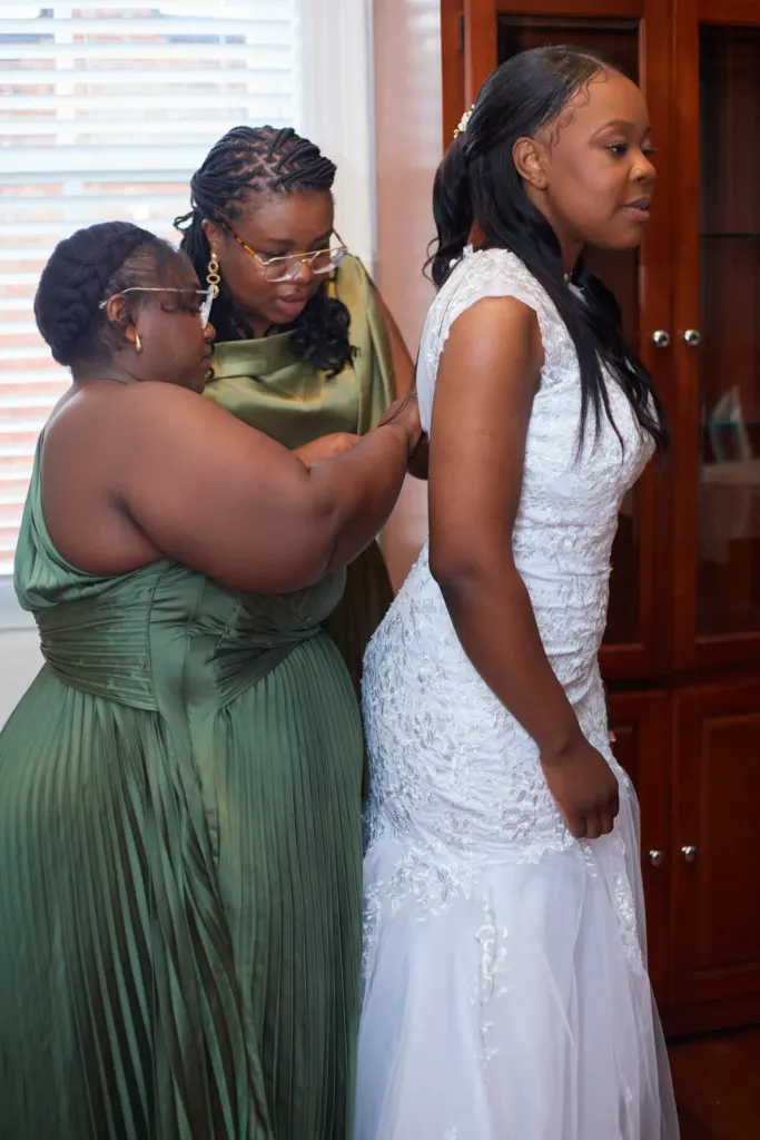 Two bridesmaids in sage satin help the bride into her lace gown by the window
