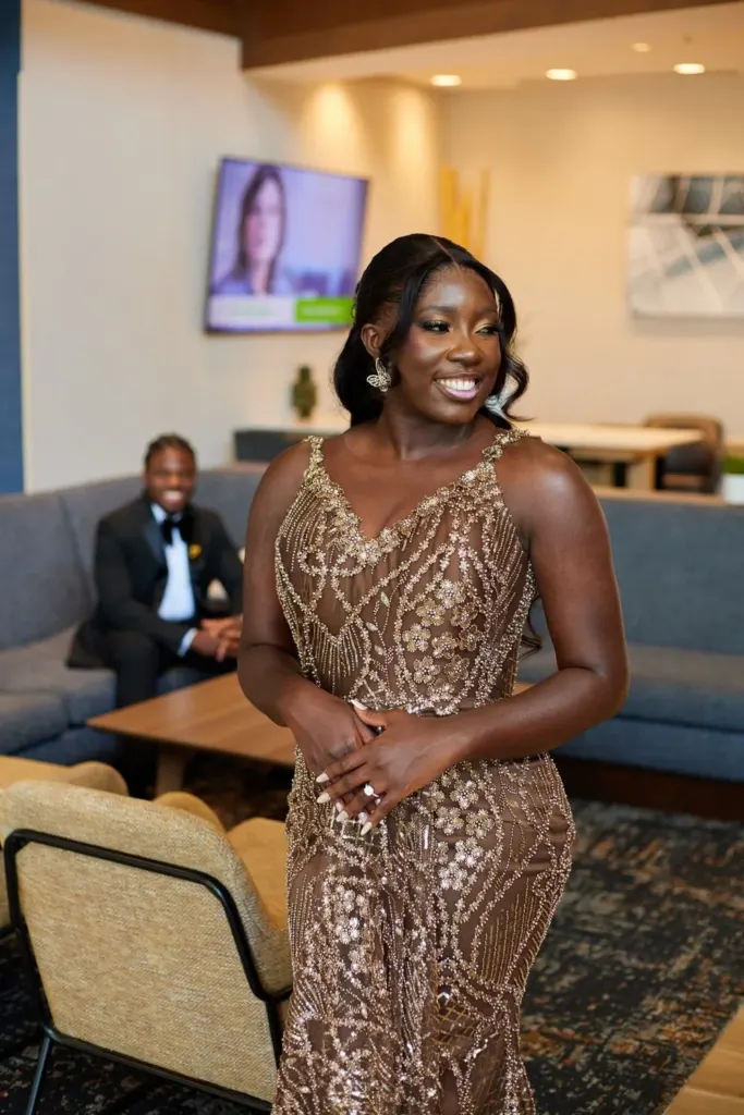 Bride in a gold beaded gown, groom watching from the sofa behind