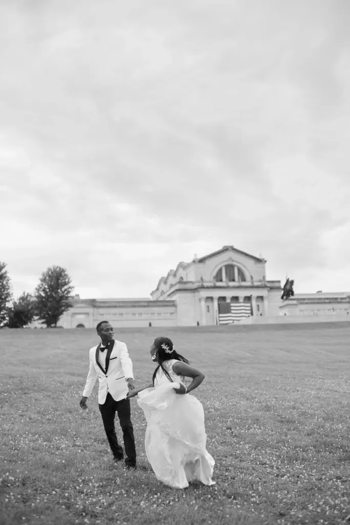 Black and white: bride gathering her gown as the couple run across a hillside lawn