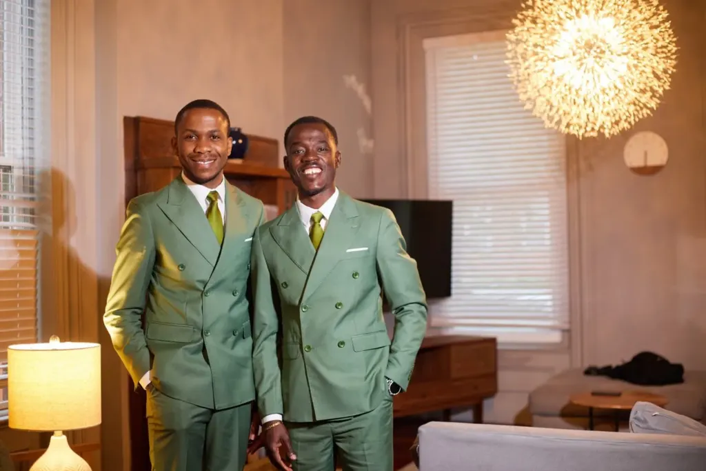 Two groomsmen in matching sage suits, laughing in warm lamplight